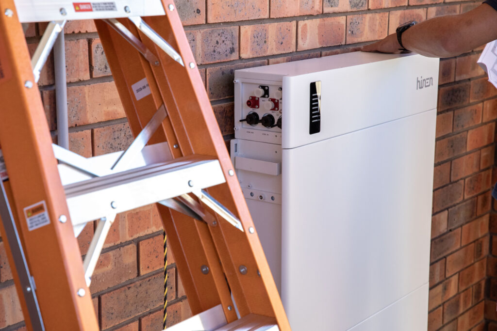 A home battery mounted against a brick wall, an installer's ladder alongside it