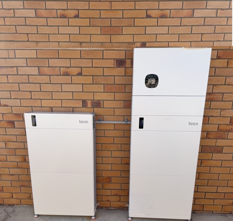 Two white home batteries of different heights against a brick wall